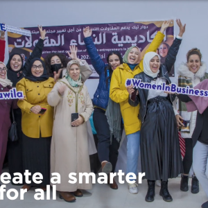A group of women, smiling, with arms raised, most wearing head-coverings. A sign in a foreign language behind them, and some holding cutout hashtag references.