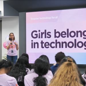 A person on stage with a microphone. "Girls belong in tech" on a large screen behind them.