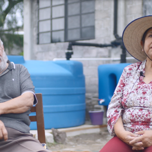 Gloria and Sergio sitting in chairs outside, in front of large water collection containers.