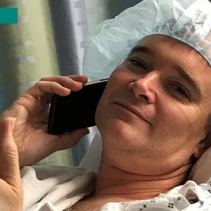 Chet Kitchen on a cell phone, wearing a hospital gown and hair covering.