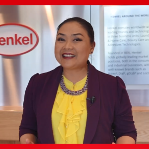  A person in front of a Henkel sign, smiling.
