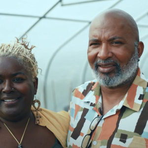 Two people standing in a greenhouse, an arm around each other.