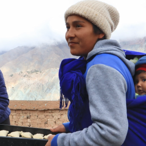 An adult carrying a baby on their pack and a tray of food in their hands, others  greet them.