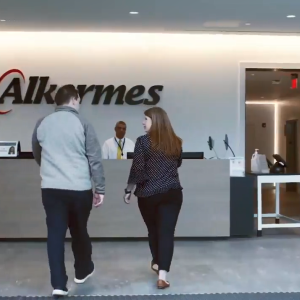 Two people approach a third at a reception desk. A sign "Alkermes" on the wall behind.