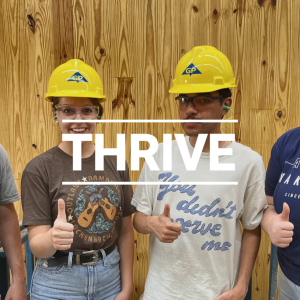 Four people giving thumbs-up, wearing matching yellow hard hats. "Thrive" on top of the image.