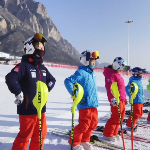 Children in a line do stretches on skis on a snowy hill.