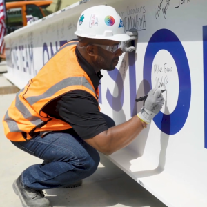 A person in hard hat and high-vis vest signs a large beam.