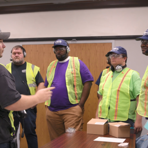 A group of people in high-vis vests gathered in a room, listening to one speaking.