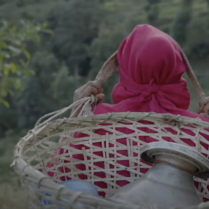 person with a pink head covering carrying a water jug in a wicker basket