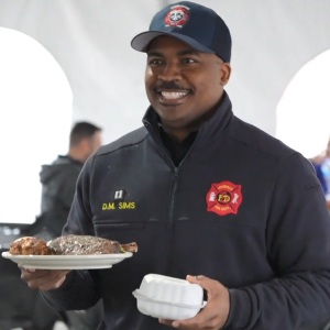 A smiling person in firefighter jacket holding a plate and container of food.