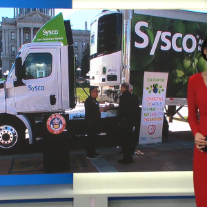 A reporter, on a screen behind them people meeting around a semi-truck outside with Sysco on the side.