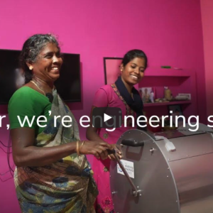 Two people with a washing machine in front of a bright pink wall