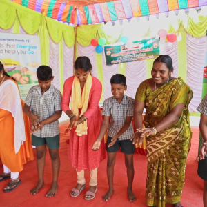A group of smiling adults and kids demonstrating hand washing.  Colorful signs behind them. 