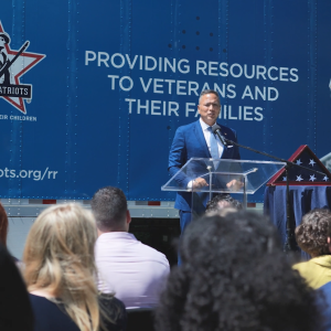 A person speaking at a podium. An american flag in a triangle box to the right and a truck behind covered with "Fallen Patriots" logos and pictures of veterans and service members.