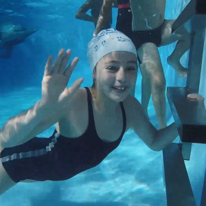 A child smiling and waving underwater in a pool, others swimming behind them.