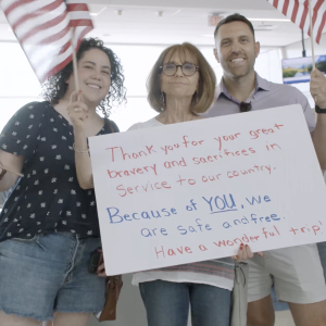 People waving American flags and holding up a sign that reads "Thank you for your great bravery and sacrifices in Service to our country. Because of YOU, We are safe and free. Have a wonder ful trip!"