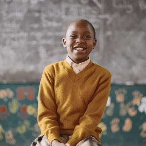 A student smiling, seated at the front of a classroom.