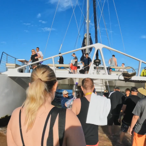 People boarding a large sail boat from a sandy shore.