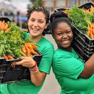 Two smiling people holding up large bins of fresh carrots.