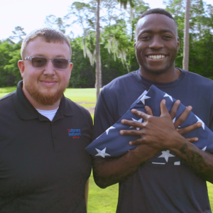 Two people posed outside. One holding a folded flag closely.