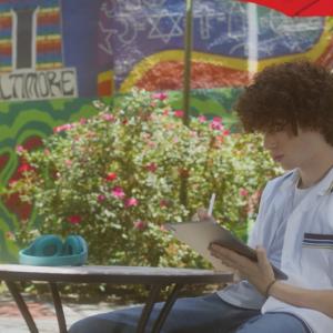 A person seated at a small table with a shade umbrella, using a tablet. A colorful mural on the wall behind them.