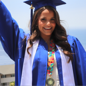 A smiling person in graduation cap and gown, one arm raised high. A scenic expanse of water behind them.
