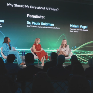Three seated speakers on a stage in front of a seated audience. Names, titles and "Why should we care about AI policy?" on a screen behind them.