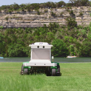 The Scythe mower in action, cutting a large grassy area. Boaters in a body of water behind it and a tall rocky ledge in the background.