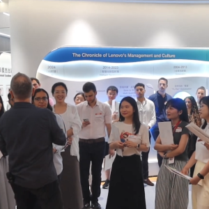 A group gathered around one speaking in an open area with displays on the wall.