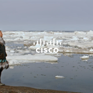 A person in warm clothing standing on a shore looking out over open water with chunks of ice and snow floating in it. "Cisco" overlaid.