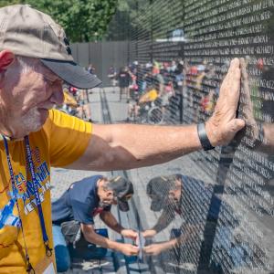 A person with a hand on a memorial wall covered in names. Their eyes are closed and head bowed.