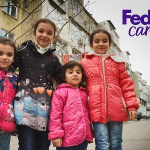 Four children posed close together, smiling outside residential buildings. "FedEx Cares" in the corner.