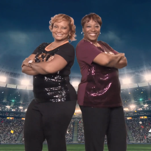 Two smiling people, leaning back to back with arms crossed. A stadium backdrop behind them.
