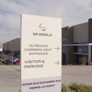 A DP World logistics facility in Perris, California, featuring a sign at the entrance directing outbound carriers and visitors to parking, with the warehouse and loading areas visible in the background under a clear sky.