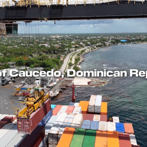 Aerial view of the Port of Caucedo in the Dominican Republic, showing container operations along the coastline with cranes lifting cargo, stacked shipping containers, and the Caribbean Sea in the background.