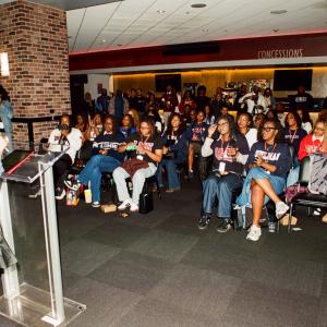 Person speaking at a podium inside an arena space while a seated audience of students listens during a program or presentation.