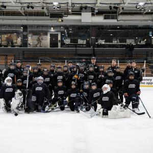 Group photo of youth participants and coaches in Black, Silver & Bold jerseys posing together on the ice rink.