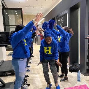 Women in blue sweatshirts holding hands up to create a tunnel for another woman in a blue sweatshirt with a yellow "H" on it to walk through