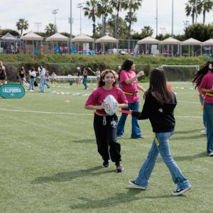 Girls wearing pink “Girls Empowerment Day” shirts play flag football on a soccer field during a youth sports clinic.