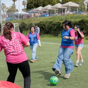 Girls wearing pink Girls Empowerment Day shirts play soccer during a youth sports clinic on an outdoor field.