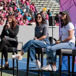 Three women participate in a panel discussion during the 5th Annual Girls Empowerment Day while a crowd of attendees sits in the stadium stands behind them.