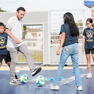 Landon Donovan coach demonstrates ball control while children in team shirts practice with multiple soccer balls on the court.