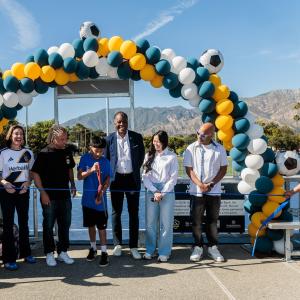 Youth participant cuts a ceremonial ribbon surrounded by LA Galaxy representatives and partners and a team mascot under a balloon arch with mountains in the background.