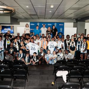 Large group of youth participants and adults pose together indoors with LA Galaxy signage and jerseys during a community event.