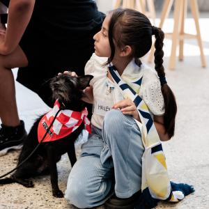 Young participant wearing a scarf sits on the floor next to a small dog in a red bandana, gently petting it.