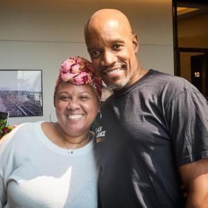 A smiling Black woman wearing a white shirt and a colorful scarf top knotted on her forehead and a smiling Black man wearing a black t-shirt