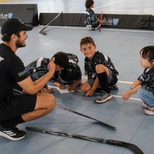 Youth participants in LA Kings ball hockey jerseys gather around a coach during a clinic session on an indoor court. Hockey sticks rest on the floor as participants listen and complete an activity together.