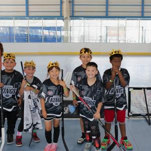 A group of youth participants wearing LA Kings ball hockey jerseys and crowns pose for a team photo on an indoor rink while holding hockey sticks and equipment.