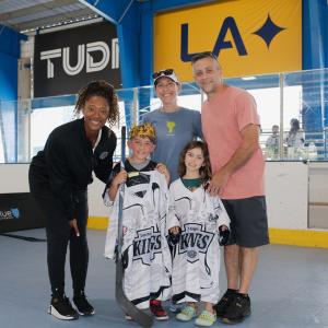 Two youth participants stand with signed LA Kings jerseys alongside family members and LA Kings Blake Bolden during a ball hockey event at an indoor sports facility.