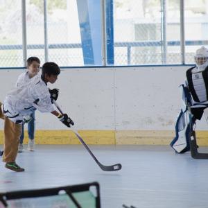 A youth participant takes a shot on goal during a ball hockey game while a goalie prepares to make a save on an indoor sports court.